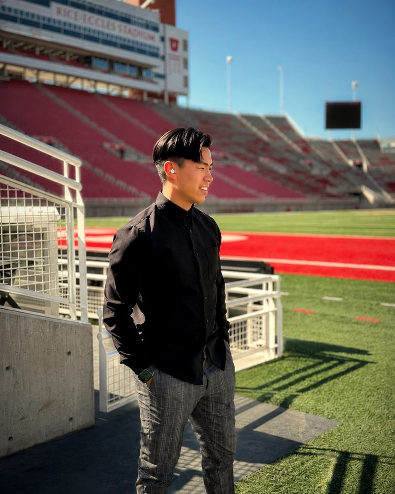 Evan Kwan smiling in a black button-up shirt on the sideline of a Salt Lake City football stadium