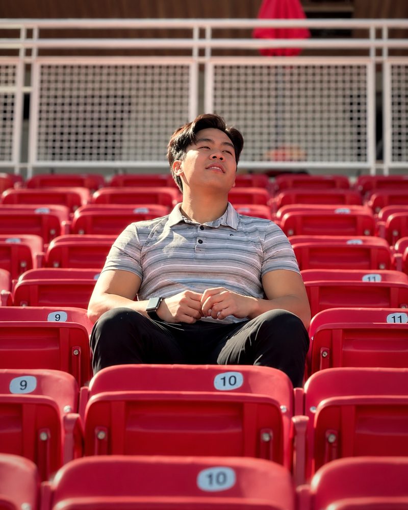 Evan Kwan sitting alone in rows of empty red stadium seats, looking up with a relaxed smile
